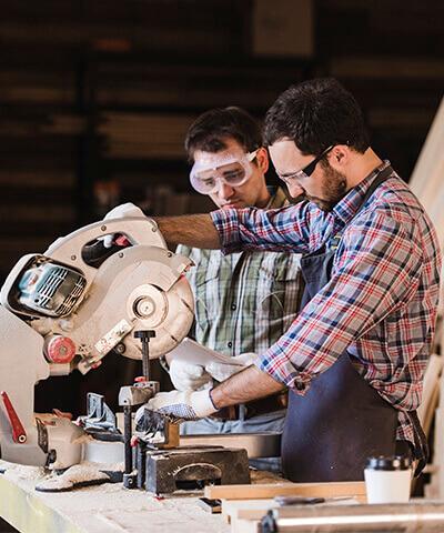 Ventajas de Seguro de Incapacidad Temporal RGA ITAutonomos - Dos hombres trabajando en una carpinteria con camisa de cuadros y gafas de proteccion, cortando madera con una sierra