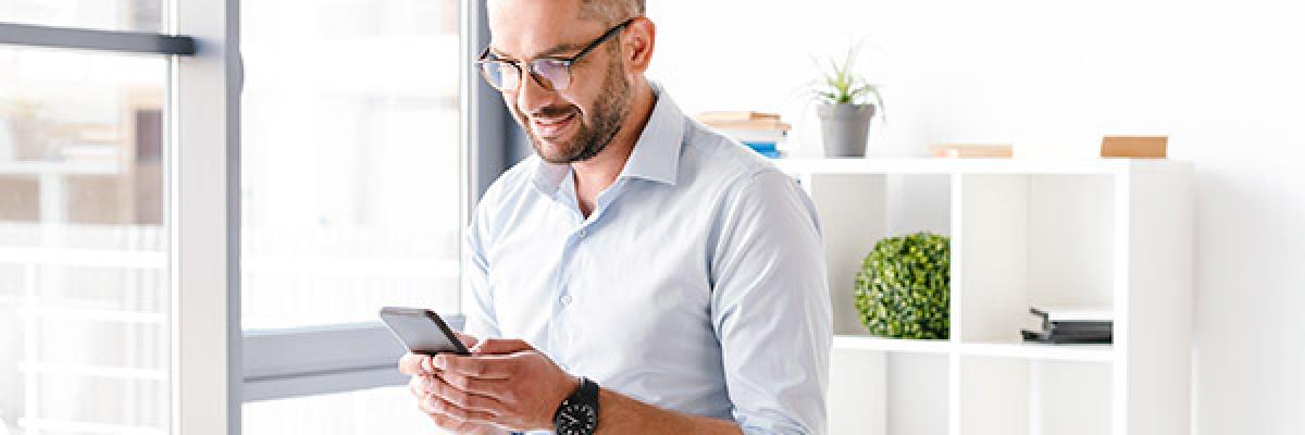 Cuenta a la Vista - Hombre de negocios con camisa y gafas sentado sobre su mesa en la oficina con un movil en la mano sonriendo